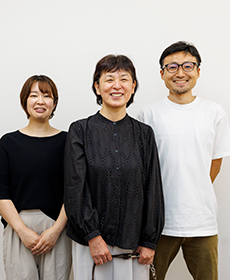 白い壁紙をバックに3名が笑顔で並んで立っている。
